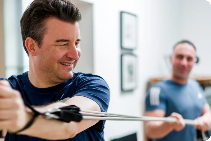 Ein Kunde trainiert lächelnd mit einem Widerstandsband, Nico Fichtler im Hintergrund